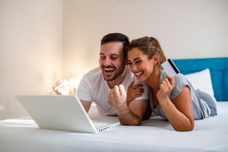 Laechelndes Paar liegt auf einem Bett, bestellt online mit Laptop und Kreditkarte – erfolgreiche Auswahl eines passenden spannbettlaken 200x200 fuer besseren Schlafkomfort