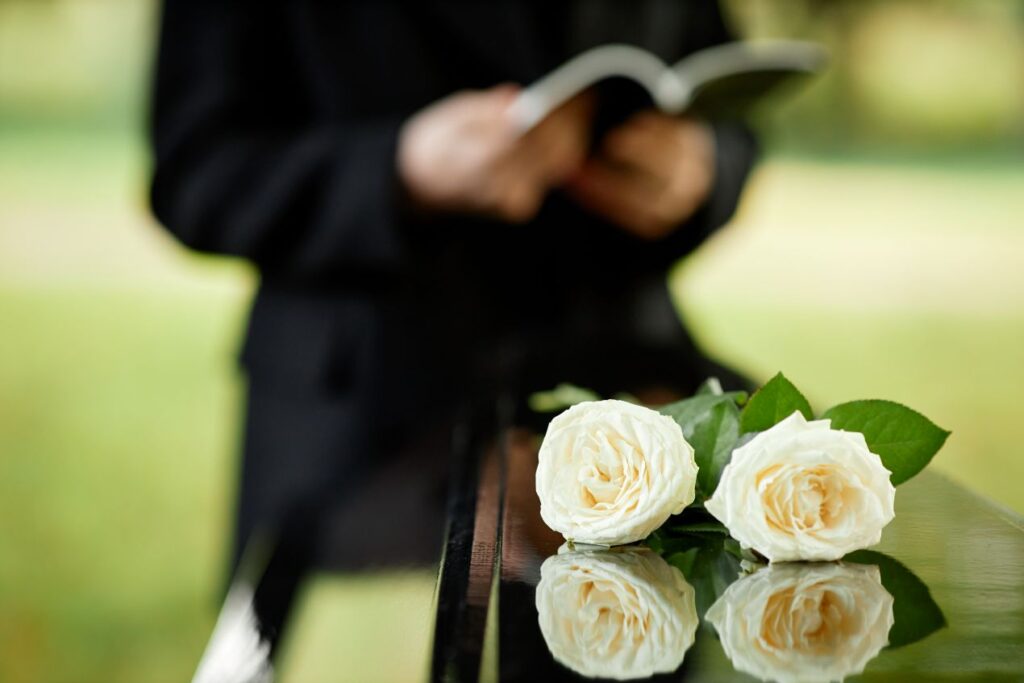 Weisse Rosen auf Sarg bei einer Beerdigung, im Hintergrund liest eine Person eine Rede – Symbolbild fuer Trauerredner werden